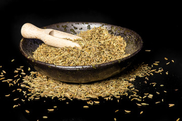 Lot of whole roman caraway seeds with a wooden scoop in a grey ceramic bowl isolated on black