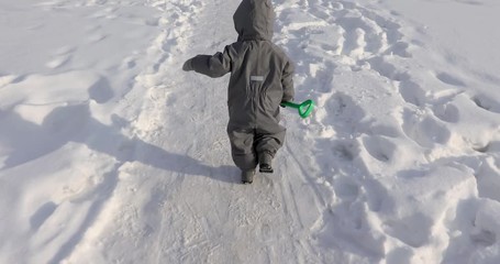 Small child runs gray jumpsuit with green shovel snow covered road