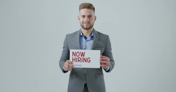 FIXED Handsome Caucasian HR manager wearing suit holding recruitment sign saying Now hiring. Light grey background. 4K UHD