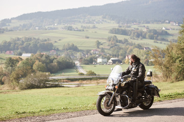 Long-haired bearded cool biker in sunglasses and black leather clothing riding cruiser powerful motorcycle along sunny asphalt road on bright summer day on background of green rural misty landscape.