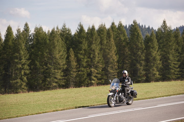 Bearded motorcyclist in helmet, sunglasses and black leather clothing riding bike down empty...