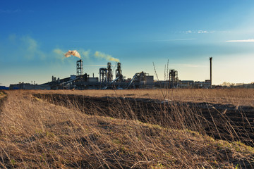 Fototapeta premium Industrial spring landscape with a view of a wood processing plant with high smoking pipes, against a background of blue sky and agricultural fields.