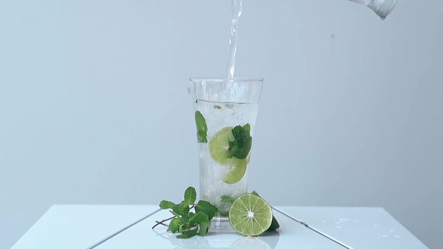 water pours into tall glass with lime pieces and mint leaves inside standing on white table against light wall