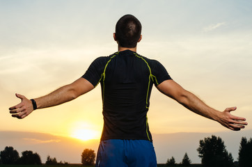 guy in sports clothing, standing looking into the sky . on the horizon the sunset. he spread his hands to meet the sun