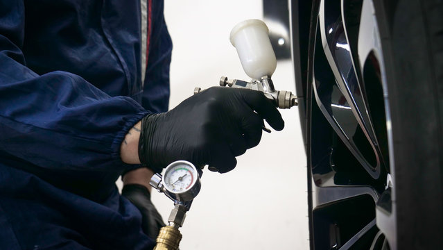 In Slow Motion A Guy (man) Professional In A Garage In Working Form With Sprays In The Hands Of A Respirator And Wearing Protective Glasses Puts Paint (ceramics) On Car Rims.