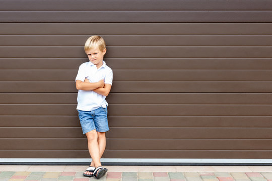 Cute grumpy blond child in casual clothing standing against brown garage door. Angry kid boy with crossed arms near house.Awkward age and parenting concept. Children and parents relations