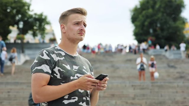 Attractive blonde guy looking around using a smartphone near huge stairway in modern city. Summertime.