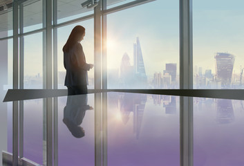 Young woman standing next to the office window with beautiful City of London view and reading...