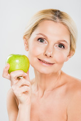 beautiful mature woman holding green apple and looking at camera on grey background