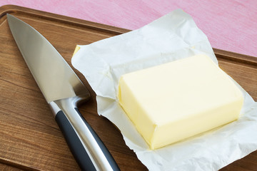 Just opened package of fresh yellow butter and a sharp kitchen knife on a brown wooden cutting board. Farmer dairy products and food ingredient.
