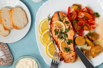 Baked salmon fillet medallion with salad of pickled vegetables and mushrooms on a white plate on a blue background, top view, flatlay