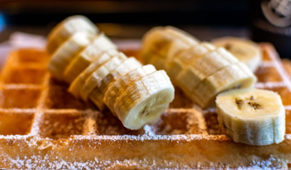Fresh square Belgian waffle with cut banana topping and sugar powder for breakfast on dark background.