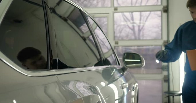 Slow Motion Man (Male) professional in working uniform and flashlight folder in his hands checks the car from all sides for serviceability Concept of: Car center, Service, Diagnostics.