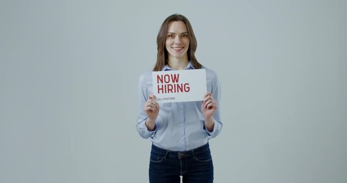 FIXED Attractive Caucasian female HR manager holding recruitment sign saying Now hiring. Light grey background. 4K UHD
