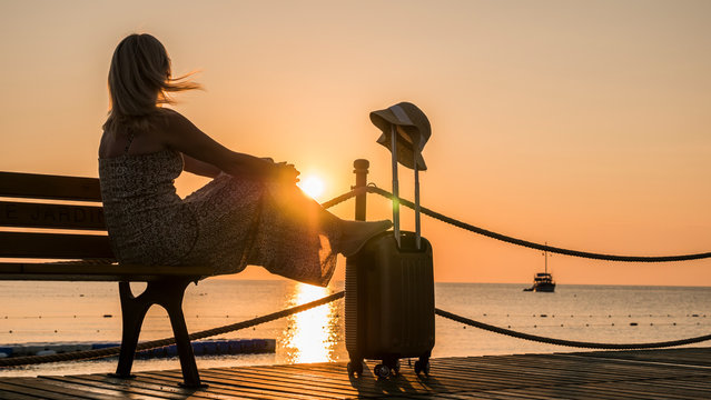 Romantic woman sitting by the sea with a travel bag at sunset. A ship is visible in the distance