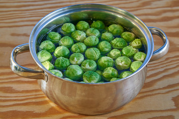 Close up picture of stainless steel cooking pot with water and bunch of fresh harvested green brussels sprouts before cooking on wooden table. Light and healthy vegetarian, vegan dish full of vitamins