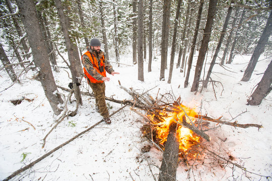 A male hunter in camouflage with a blaze orange vest on warms his hands by a fire he started when the weather turned colder during an elk and deer hunting trip in Northwest Montana.