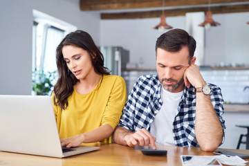 Worried couple sitting at dining table, using laptop, checking finances