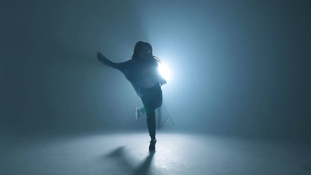 Young woman silhouette dressed in street fashion wear dancing hip-hop style over studio blue light background with flare effects, slow motion