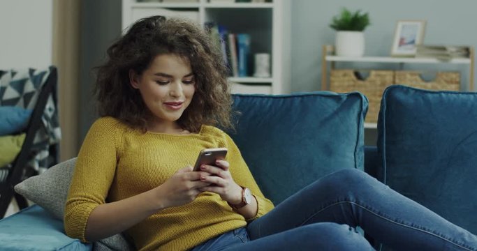 Young cheerful Caucasian beautiful curly woman scrolling or typing on the smartphone while lying on the sofa at home.