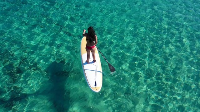 Aerial drone bird's eye view video of attractive fit young woman practicing stand up paddle board or SUP in tropical caribbean sapphire crystal clear sandy exotic beach