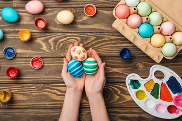 top view of woman holding painted easter eggs at wooden table with paints and color palette
