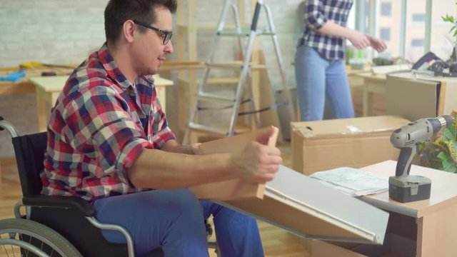 Disabled person in a wheelchair collects a nightstand in a new modern apartment next to his wife