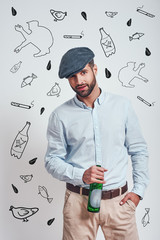 Bad boy. Bearded brutal man in hat holding a beer and looking ar camera while standing against grey background with hand drawn doodles on it.