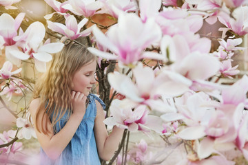 Beautiful little blond girl in blue dress holding flowers of magnolia under blossom magnolia tree. 