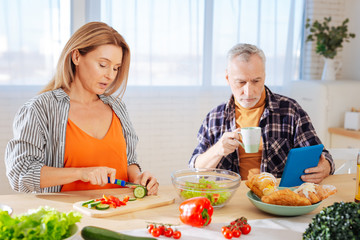 Bearded husband reading news on tablet while wife cooking salad