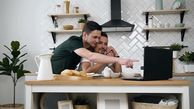 Lovely Caucasian Gay Couple, Homosexual Couple, Are Hugging And Having Breakfast At The Dinner Table Using Internet On Laptop In Kitchen At Home. Lifestyle Of Gay Couple