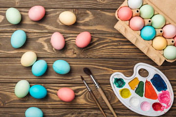 top view of pastel easter eggs in cardboard box with color palette and paintbrushes on wooden table