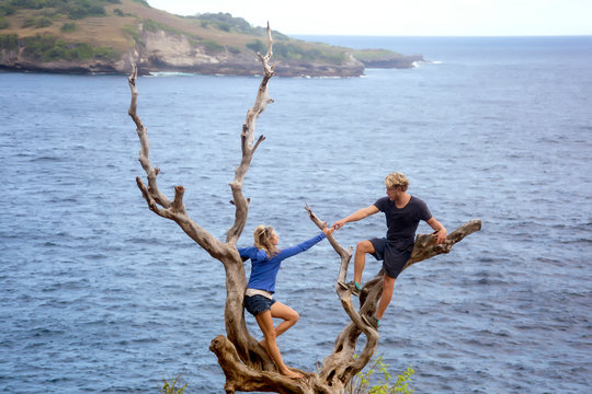 Couple climbing on bare tree on coastline, Nusa Penida, Bali, Indonesia