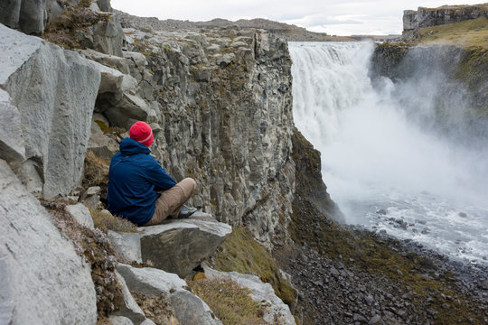 Young man sitting down looking at Detifoss waterfall in Iceland.
