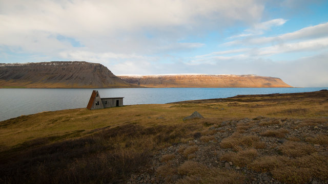 A lone A-frame tiny house sits over a fjord in a vast landscape in Iceland.