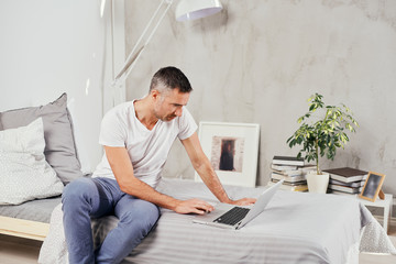 Handsome Caucasian middle aged man dressed casual using laptop while sitting on bed in bedroom.