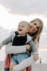 Fototapeta na wymiar Adorably Perfect Young Mother and Baby Toddler Son Family Laughing, Smiling, and Holding One Another at the Sandy Beach During Sunset Outside By the Ocean Water on Vacation