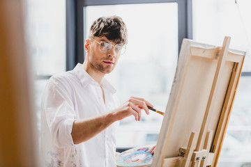 Naklejka na ściany i meble selective focus of handsome artist in glasses looking at camera while drawing on canvas