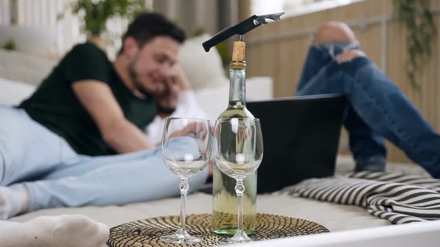 Bottle Wine With Two Glass Standing On Lovely Gay Couple Bed. Attractive Gay Couple Using Laptop Together With Funny Emotion Laying In The Bed At Bedroom. Gay Men Spending Time Together