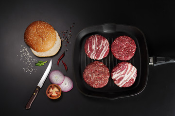 Cutlets for burgers in a grill pan and other ingredients on a black background, top view.