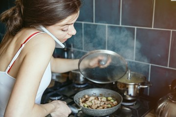 A young woman is cooking the dish while talking on the phone. She is in the kitchen room. Working mother concept.
