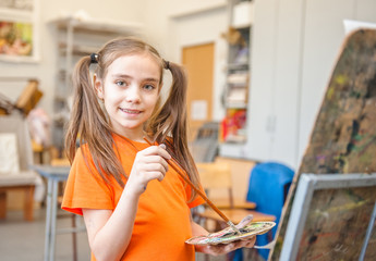 Portrait of a lovely little girl painting a picture in a studio or art school. Creative pensive painter child paints a colorful picture on canvas in workshop. Talented kids