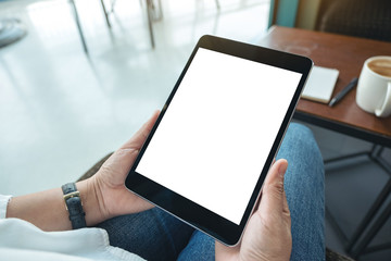 Mockup image of a woman sitting and holding black tablet pc with blank white desktop screen in cafe