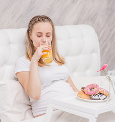 Young pregnant woman having breakfast in bed with juice and donuts