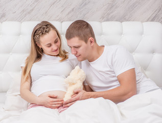 Happy couple waiting for the baby. Young family on the bed with toy bear on the belly of the pregnant mother