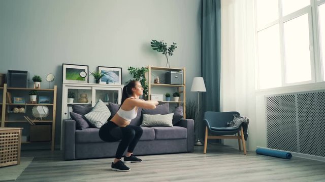 Strong girl student is squatting at home standing on floor of beautiful light flat focused on exercise. Woman is wearing trendy leggings, short top and sneakers.