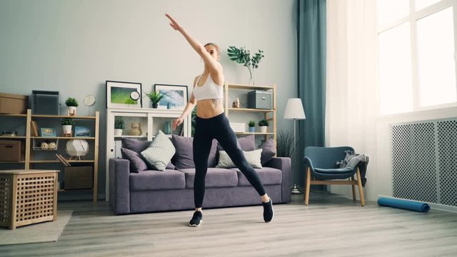 Fit girl is practising at home squatting and raising arms exercising on floor in light living room. Modern young people, indoor sports and apartments concept.