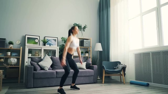 Active young lady is concentrated on sports exercises squatting and jumping on floor of light apartment enjoying movement and activity. Millennails and modern lifestyle concept.