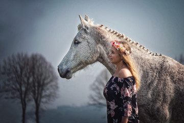 Portrait Frau und weißes Pferd Schimmel in Blumenkleid mit Blumenkranz im Haar im Wald in der Natur mit Himmel im Hintergrund