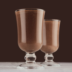 Winter hot drink cocoa in two elegant glass on dark brown wall, white wood table, square.